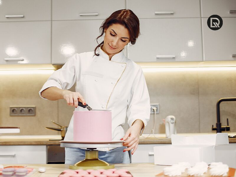 Mulher usando avental e segurando um saco de confeiteiro, decorando um bolo em uma cozinha.